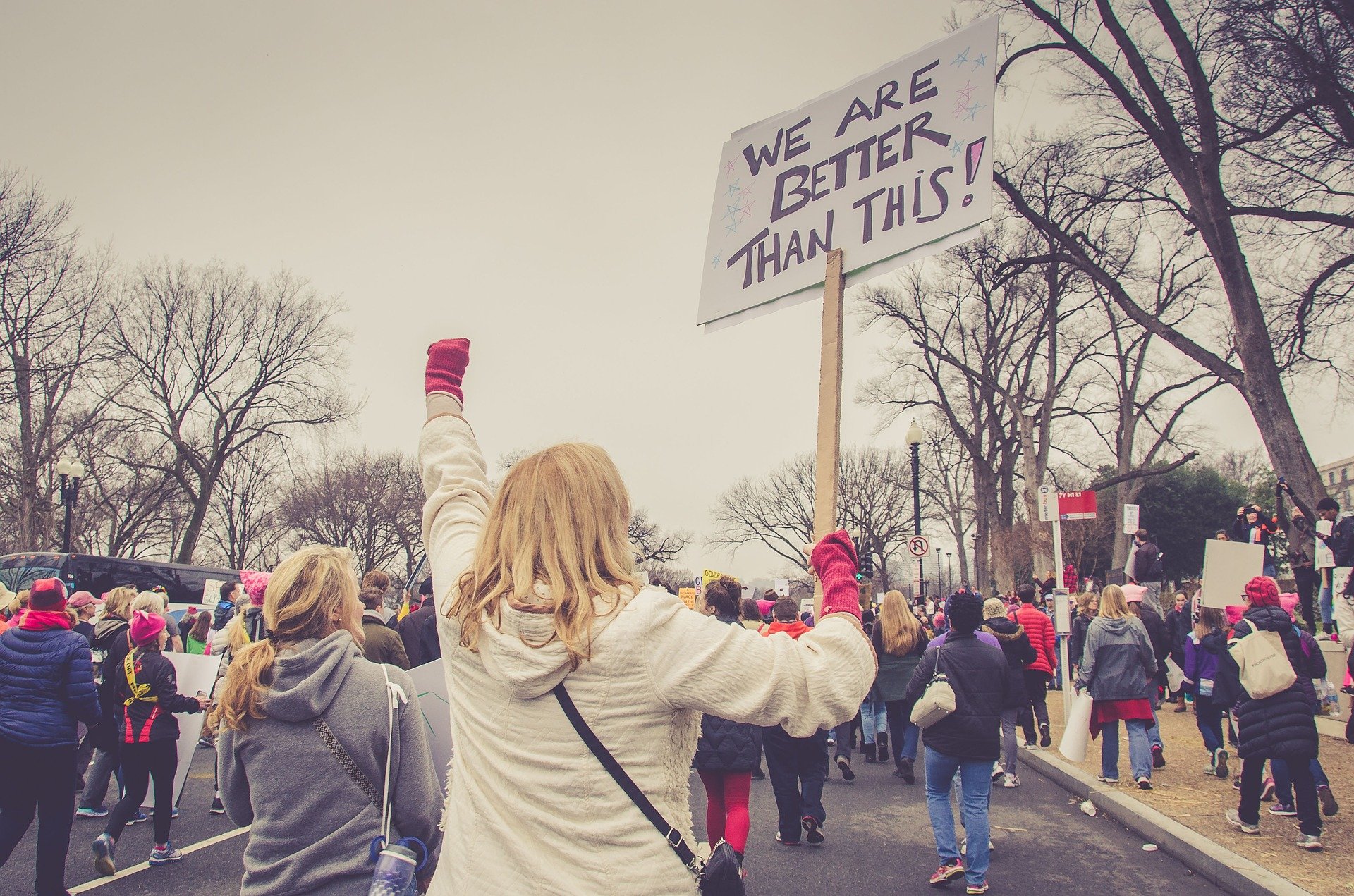 Woman with blond hair holding sign that reads "We are better than this!" at a gathering